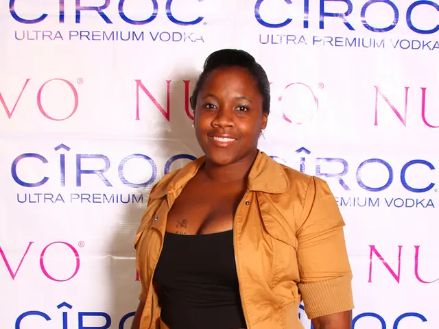 In this image there is a lady standing with a smile on her face, behind her there is a wall with...